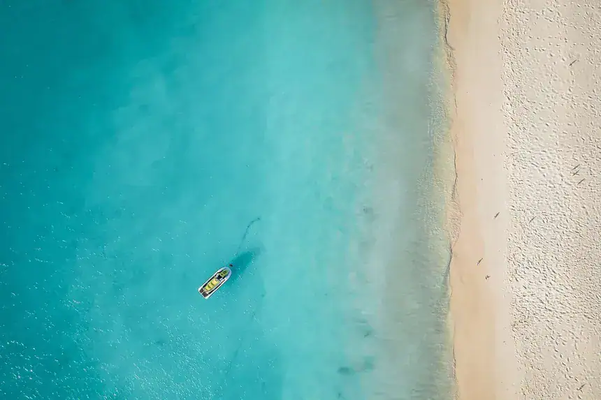 Aerial view of a small boat floating on turquoise waters near the white sandy shore of Eagle Beach in Aruba.