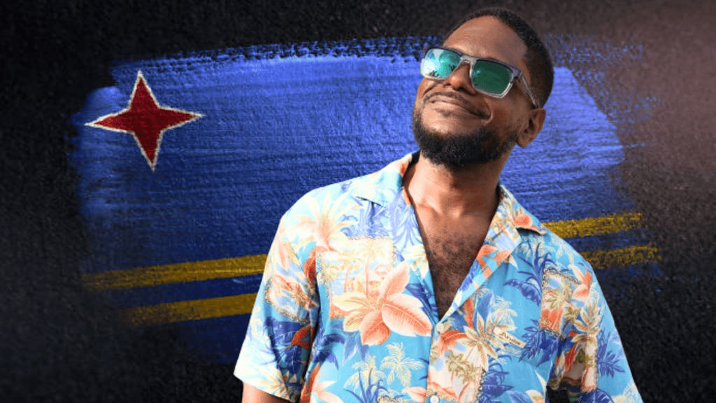Beach Lama in floral shirt standing in front of a painted Aruban flag with red star and yellow stripes.