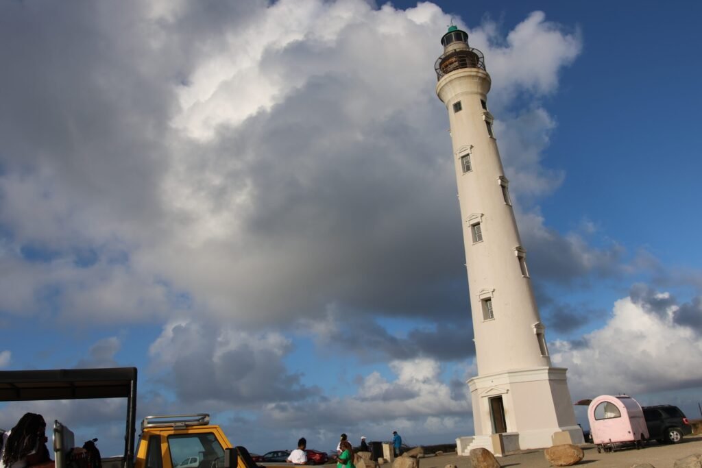 The California Lighthouse in Aruba captured from a distance with people and vehicles at its base under a cloudy sky.