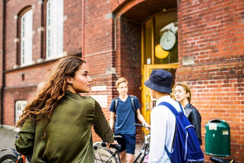 School students standing and talking outside brick school building with bicycles in the background, young people, education, student life, peer interaction, urban school setting.