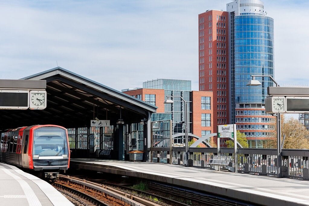 Modern urban train station with contemporary architecture and city backdrop, featuring a red train ready for departure, clocks, and clear signage, in a vibrant cityscape.