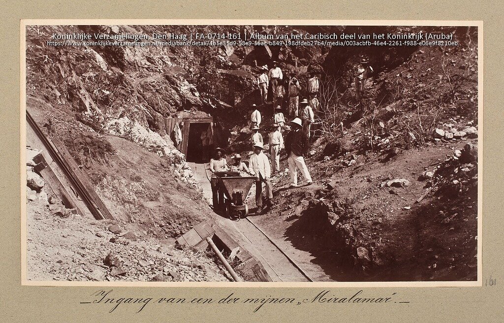 Vintage photo of miners working at Miralamar gold mine in Aruba, showing early mining techniques on a rugged hillside with a group of workers and equipment.